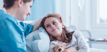 A smiling healthcare worker comforting a young girl in a hospital bed holding a teddy bear.